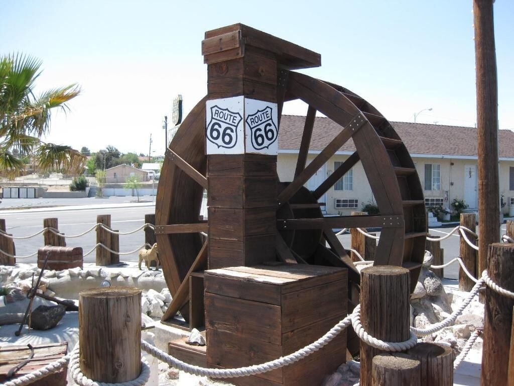 A wooden water wheel with Route 66 signs, surrounded by wooden posts and rope barriers.