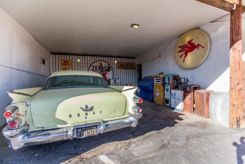 Un coche antiguo estacionado en un garaje con un letrero de Texaco, el logotipo de un gallo rojo y electrodomésticos antiguos.