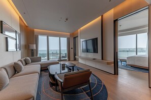 A modern hotel room with a large sofa, a TV, a balcony with a view, and a dining area.