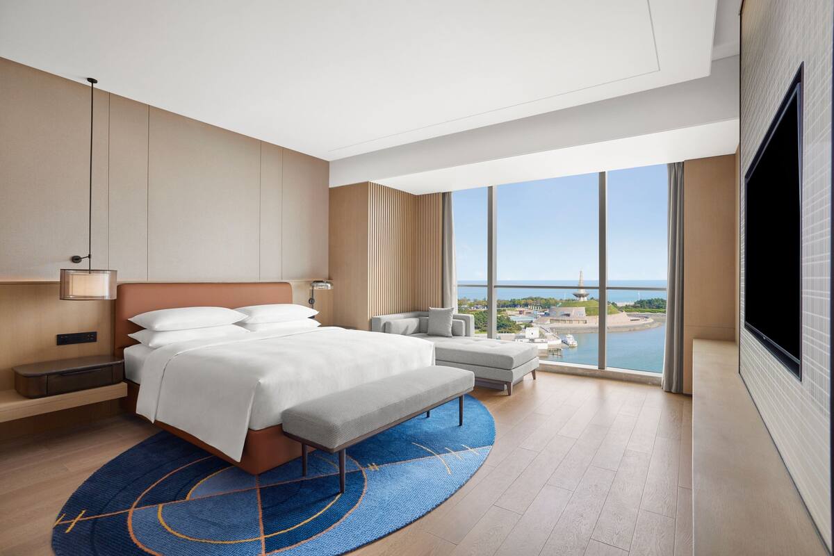 A modern hotel room with a large bed, a seating area, and a view of the sea.