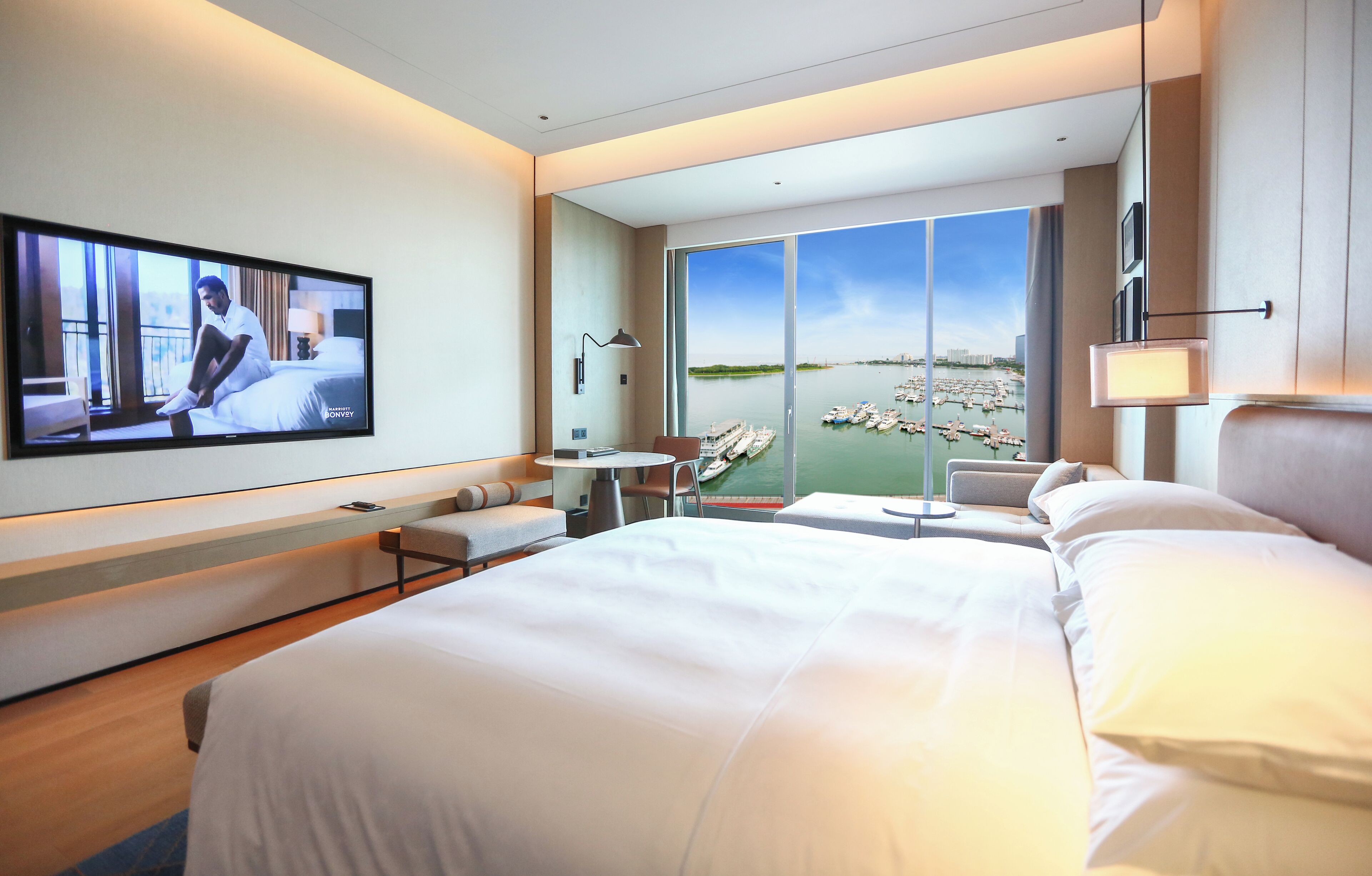 A modern hotel room with a large bed, a TV on the wall, a desk with a chair, and a view of the marina through large windows.