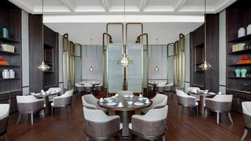 A spacious dining area with round tables and modern chairs, featuring a central chandelier and mirrored walls.