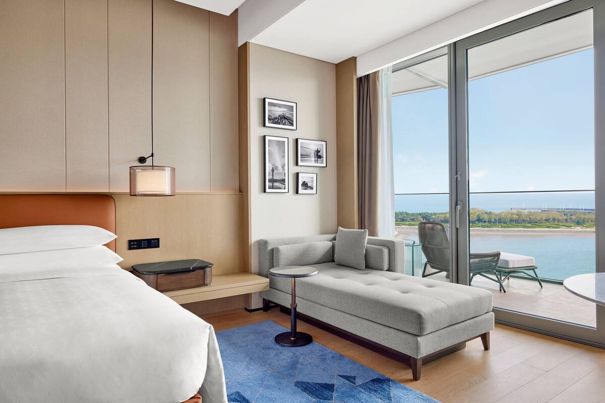 A modern hotel room with a large bed, a sofa, a small table, and a balcony with a view of the water.