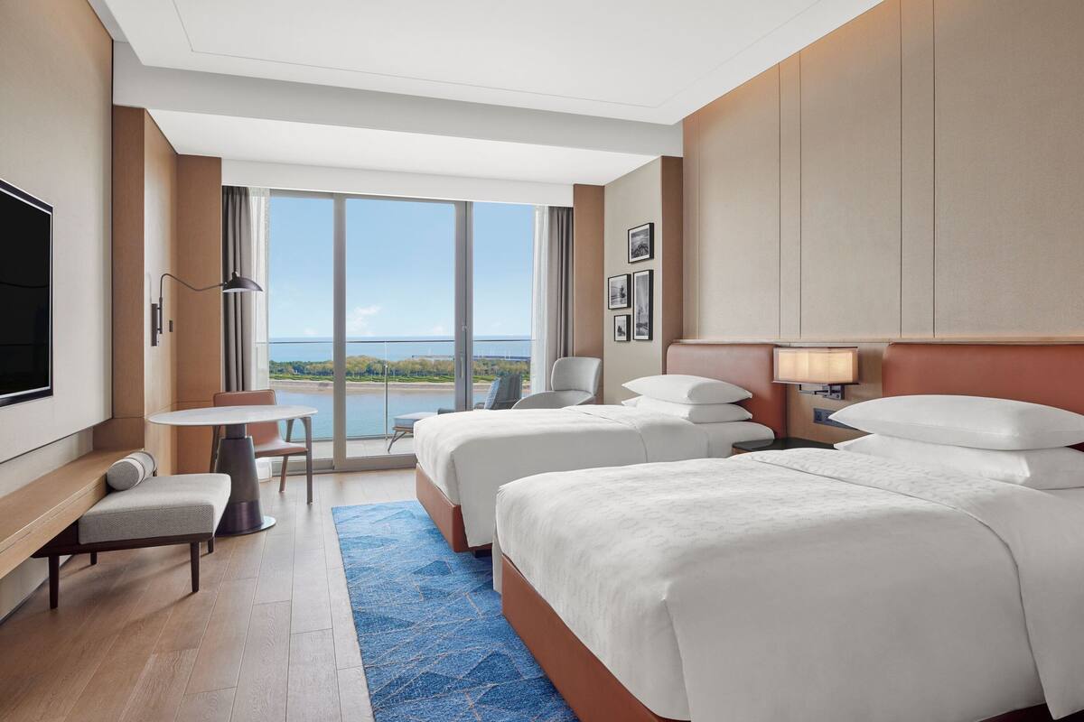 A hotel room with two beds, a TV, a desk, and a large window offering a view of the sea.