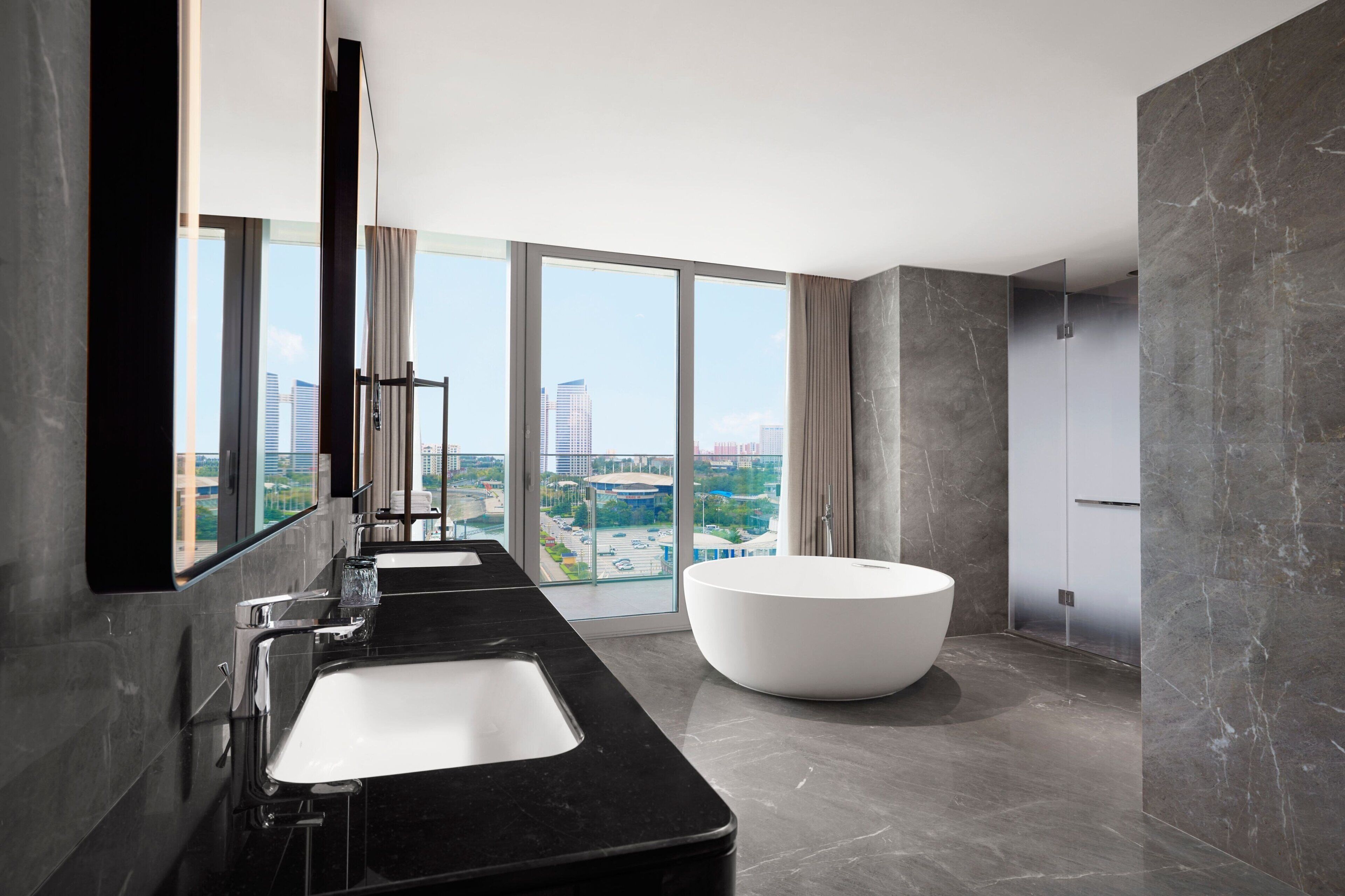 A modern bathroom with a large bathtub, two sinks, and a large window offering a city view.