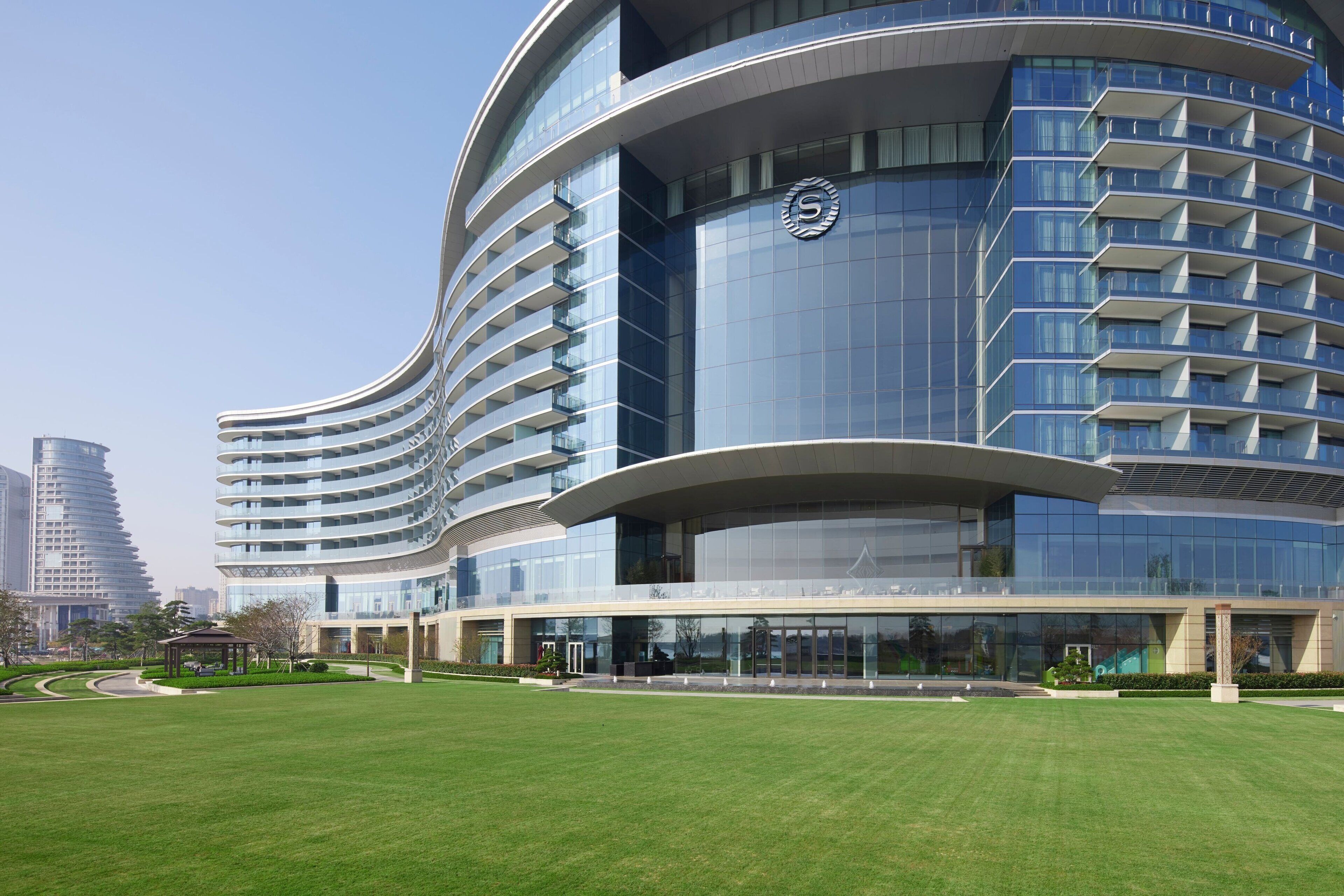 A modern hotel with a curved facade, multiple balconies, and a well-maintained lawn.