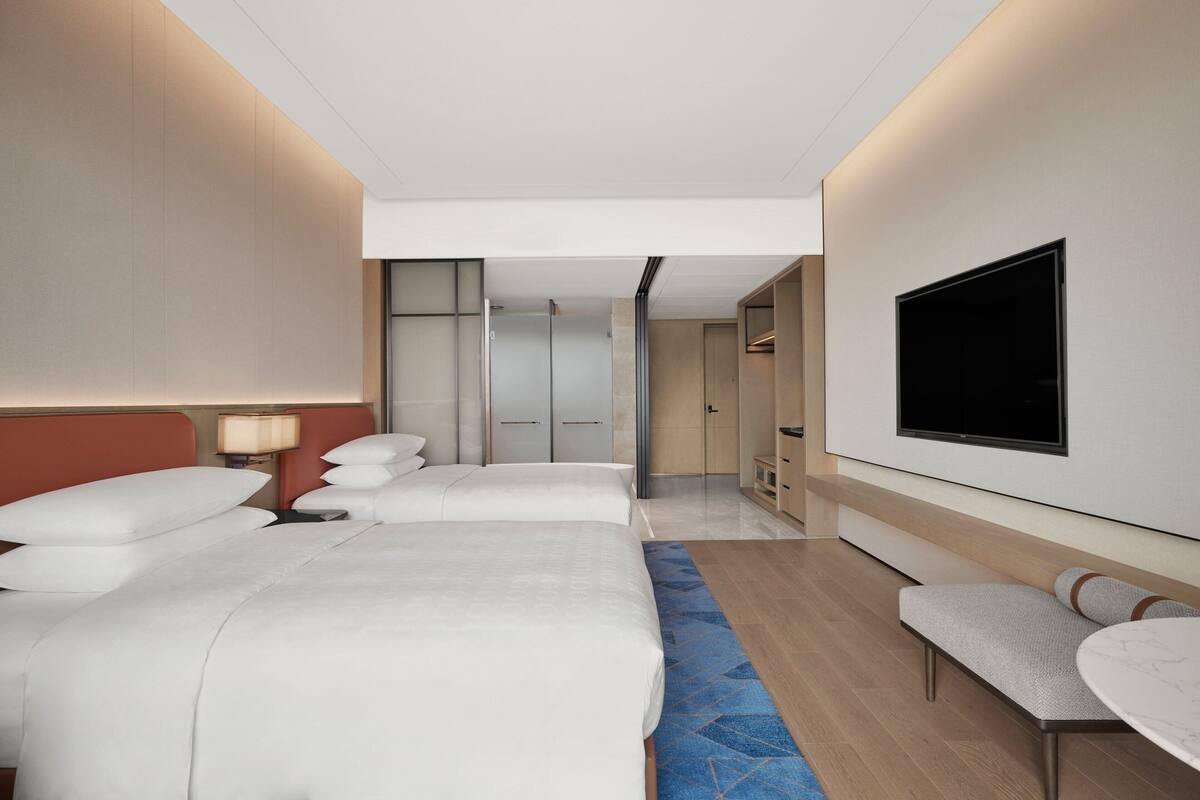 A modern hotel room with two beds, a TV, a seating area, and a bathroom visible through a glass door.