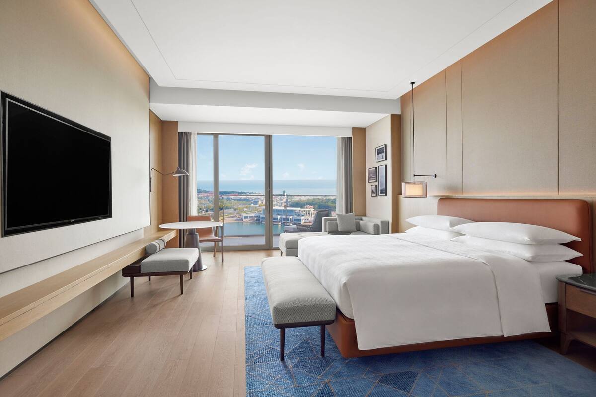 A modern hotel room with a large bed, a flat-screen TV, a seating area with a table and chairs, and a view of the city and sea through large windows.