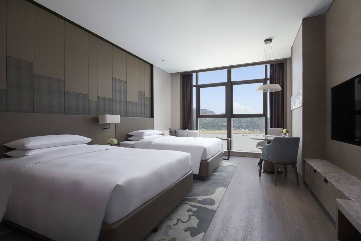 A hotel room with two beds, a desk, a chair, a TV, and a large window offering a view.