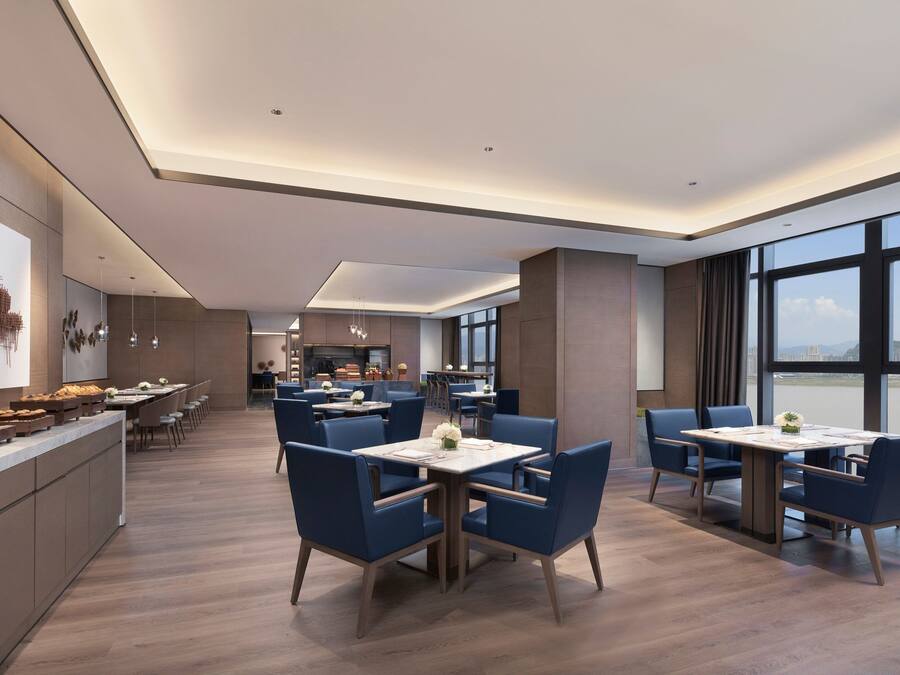 Un comedor moderno de hotel con varias mesas y sillas, una cocina al fondo y amplios ventanales que ofrecen una vista.