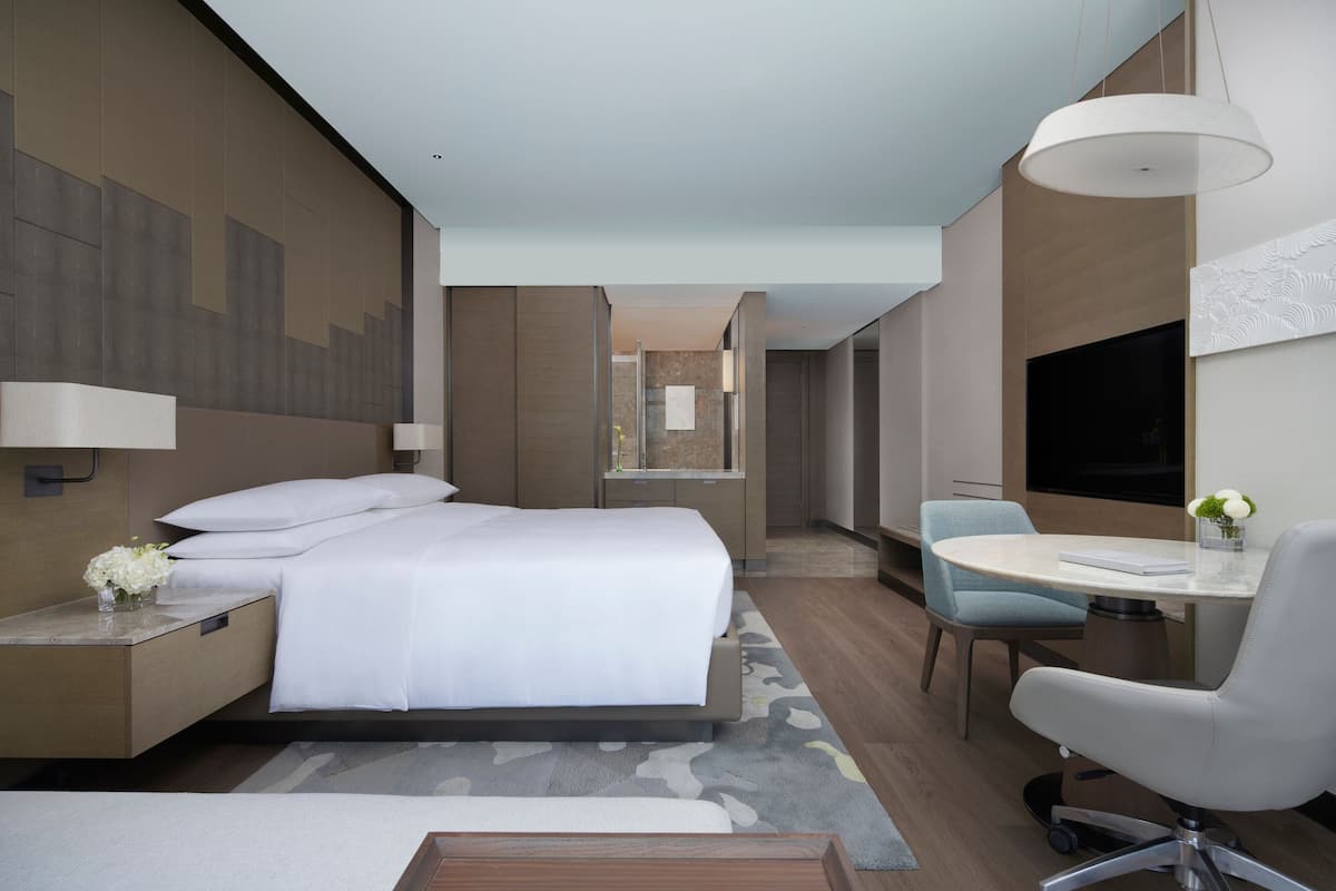 A modern hotel room with a large bed, a desk with a chair, a TV, and a bathroom visible in the background.