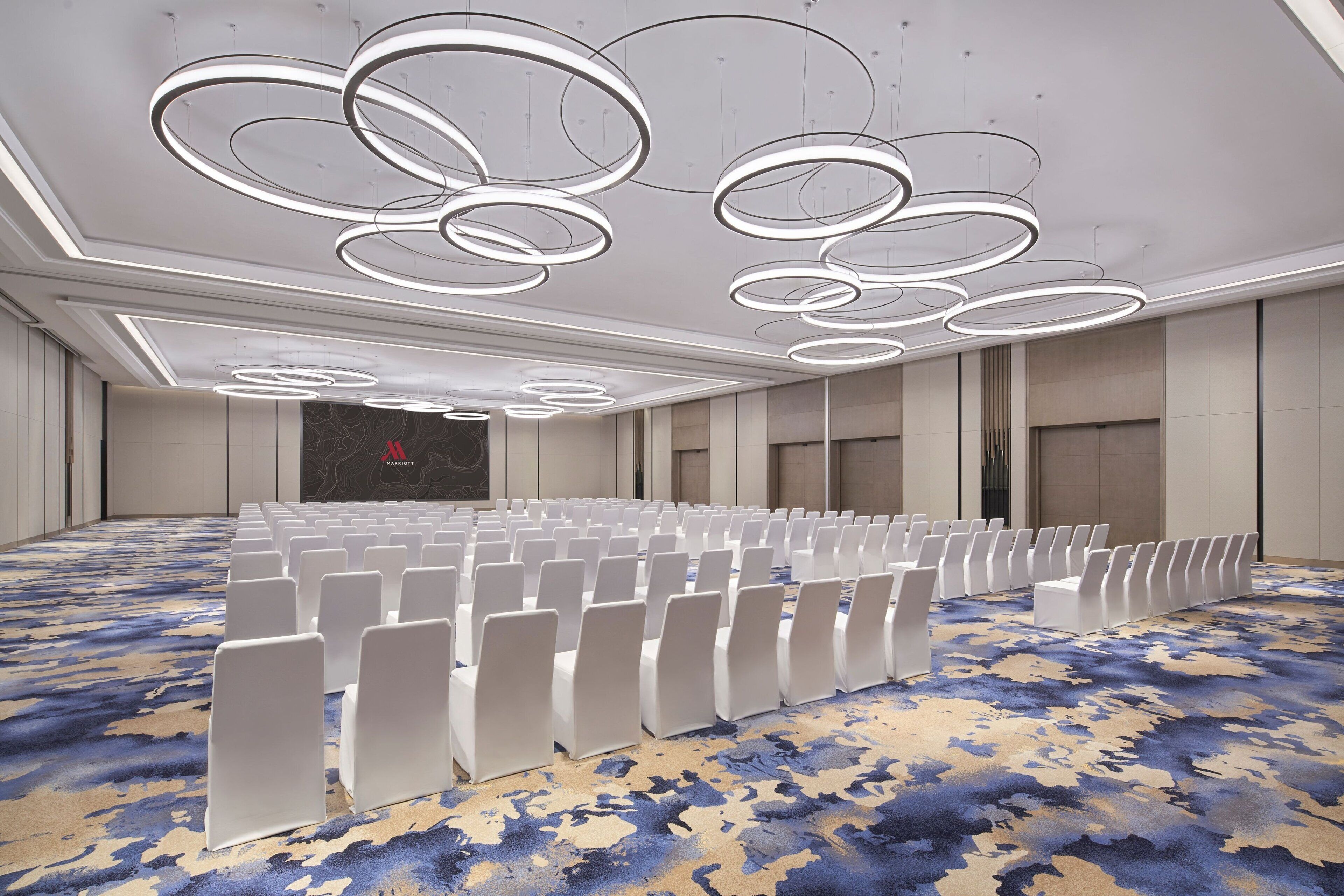 A conference room with rows of white chairs, a patterned carpet, and modern circular ceiling lights.