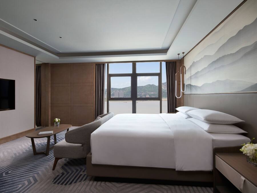 Ein modernes Hotelzimmer mit einem großen Bett, einem Flachbildfernseher, einem kleinen Tisch und Blick auf die Stadt durch große Fenster.