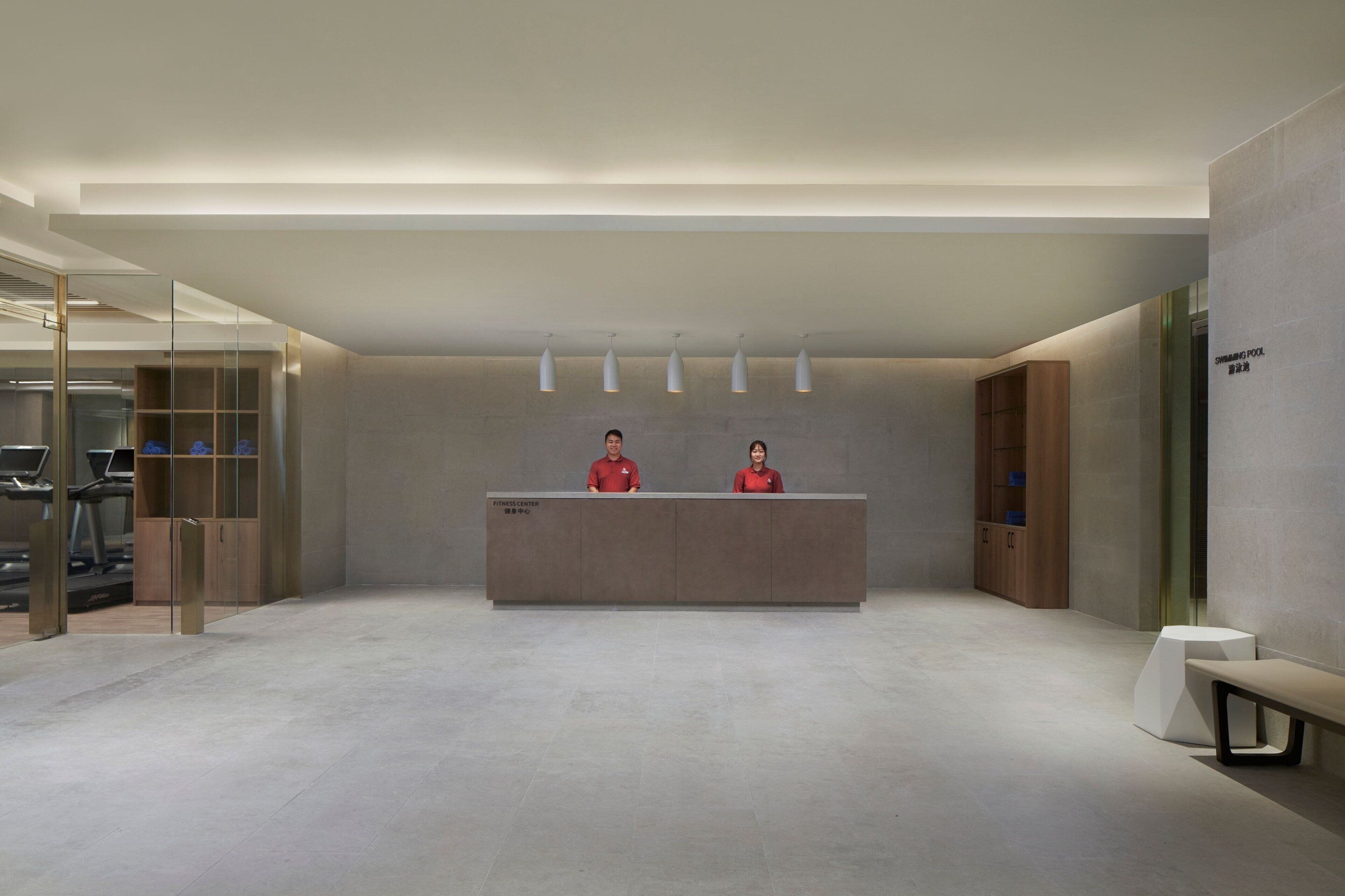 A reception area with a reception desk, two staff members in red uniforms, a glass partition, and a treadmill in the background.