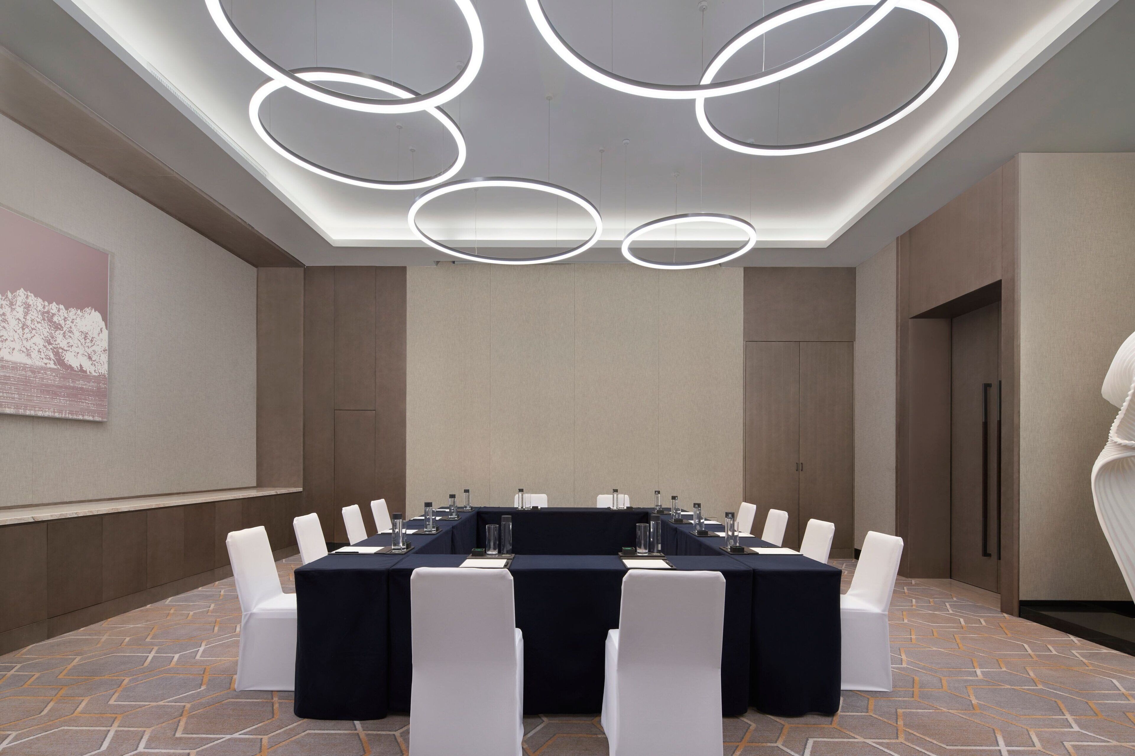 Een vergaderzaal met een lange tafel, witte stoelen en een moderne plafondlamp.