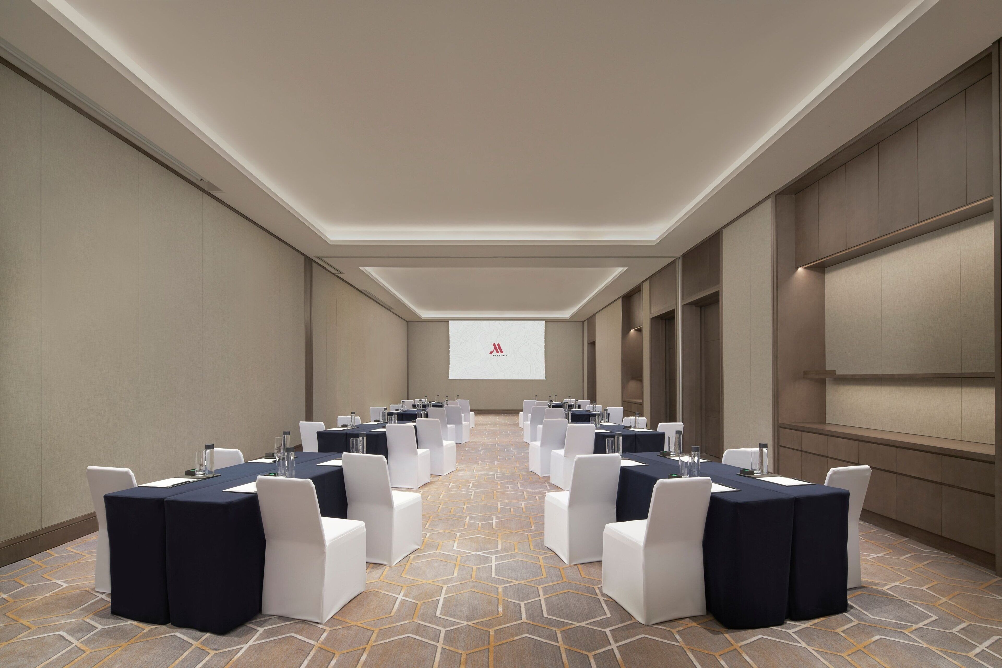Un salón de conferencias con una larga mesa, sillas blancas y una pantalla de proyector al frente.