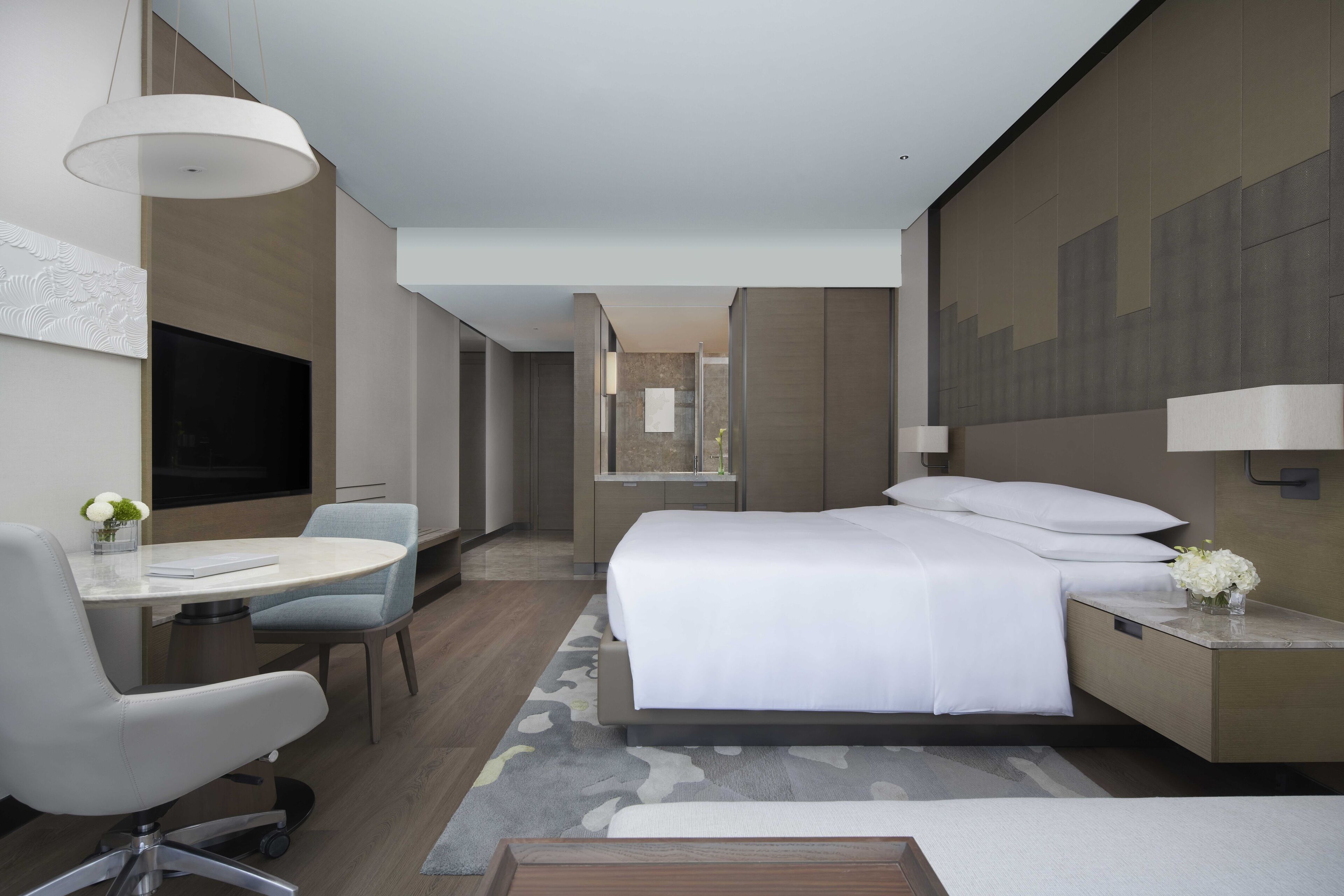 A modern hotel room with a bed, bedside table, chair, desk, and a TV.