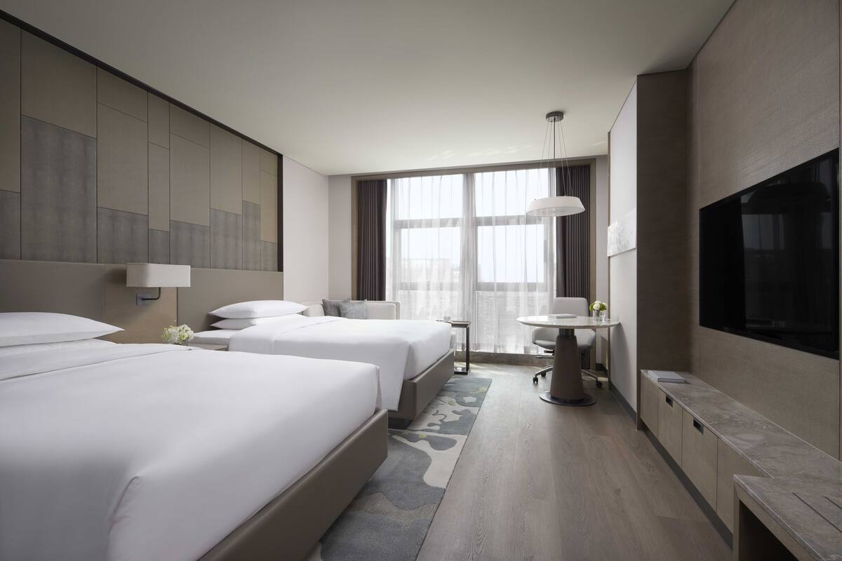 A modern hotel room with two beds, a desk, a TV, and large windows.