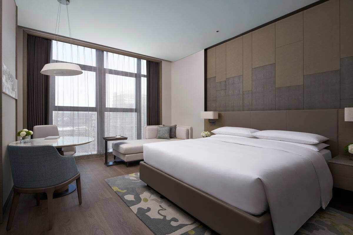 A modern hotel room with a large bed, a seating area with a table and chairs, and a large window with curtains.