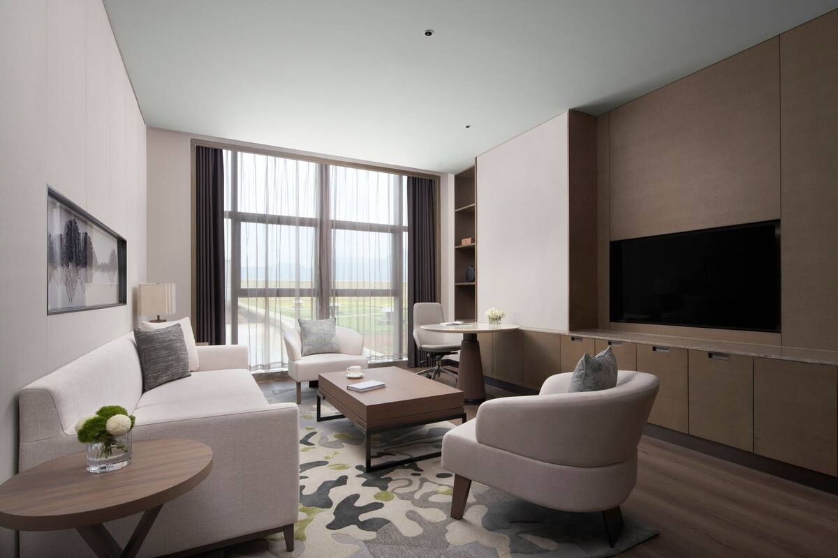A modern hotel room with a sofa, armchair, coffee table, and a flat-screen TV mounted on the wall.