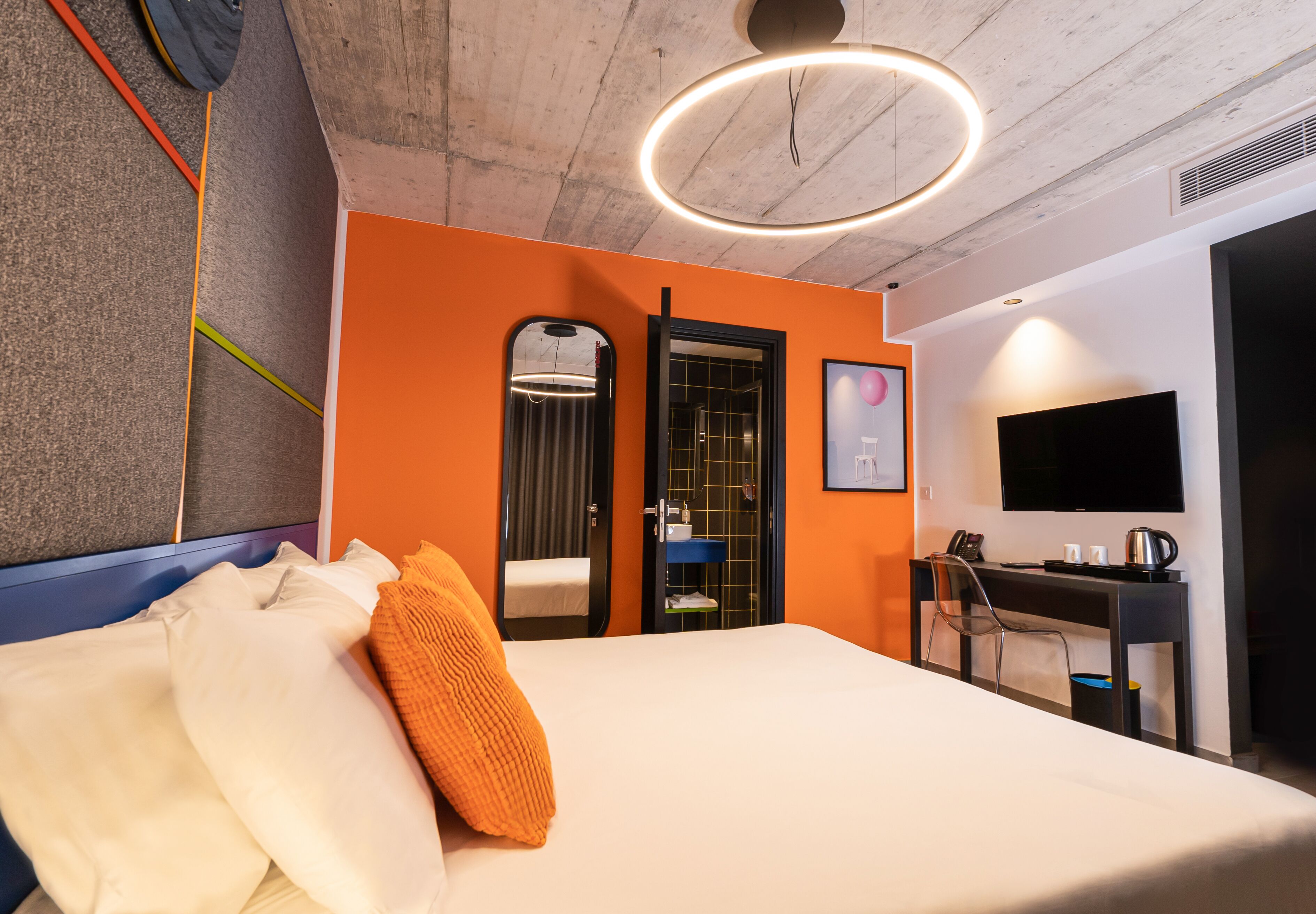Quarto de hotel com cama, escrivaninha, cadeira, televisão e banheiro com espelho e chuveiro.