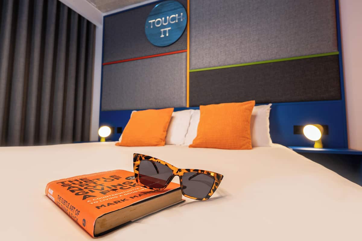 A neatly made bed with orange pillows, a book, and sunglasses on it.