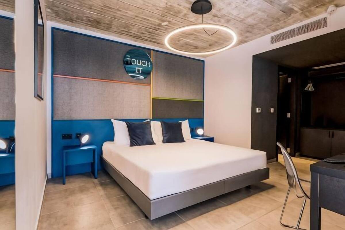 A modern hotel room with a large bed, bedside tables, a wall-mounted TV, and a visible door leading to another room.