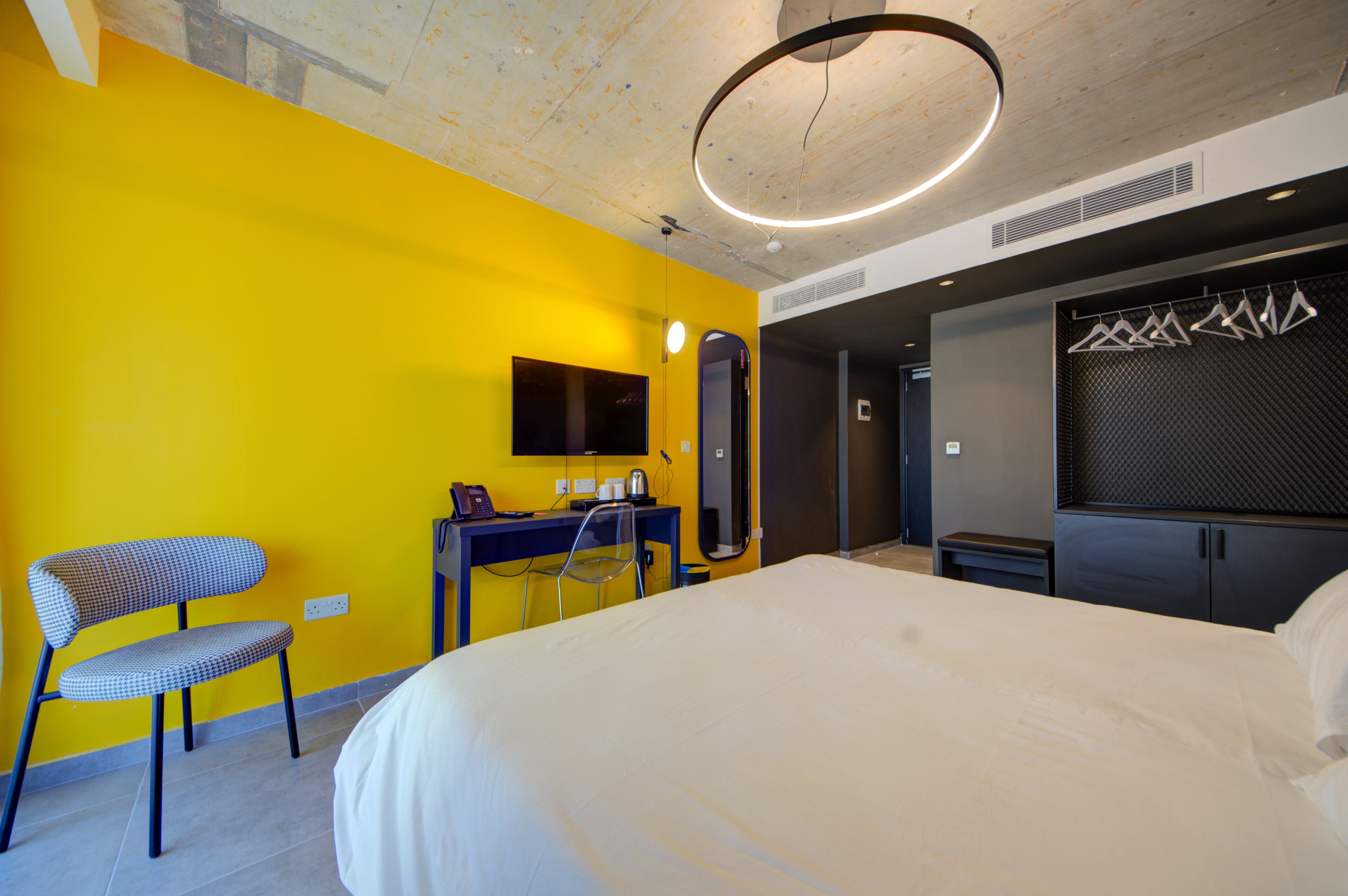 A hotel room with a large bed, a desk with a chair, a flat-screen TV mounted on the wall, and a modern pendant light.