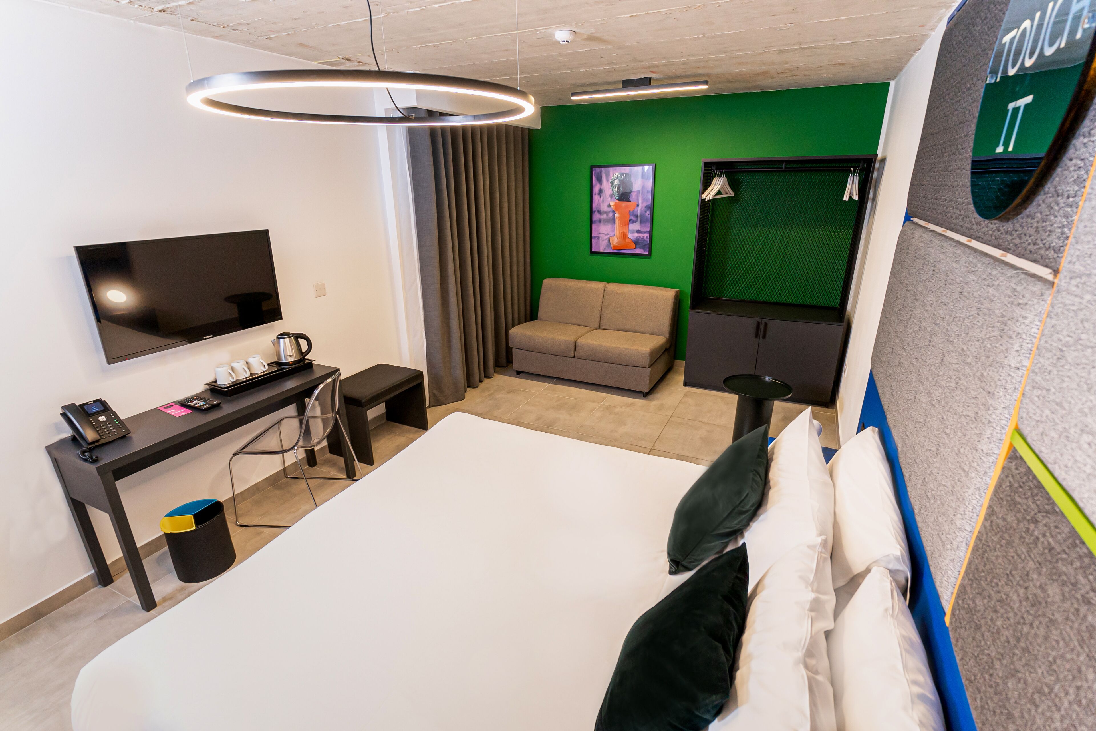 A modern hotel room with a large bed, a desk with a telephone, a flat-screen TV, and a green accent wall.