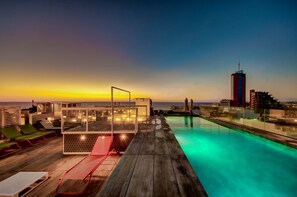 Zona de piscina en la azotea con sillones de descanso, cerramiento de vidrio y vistas del horizonte urbano al atardecer.