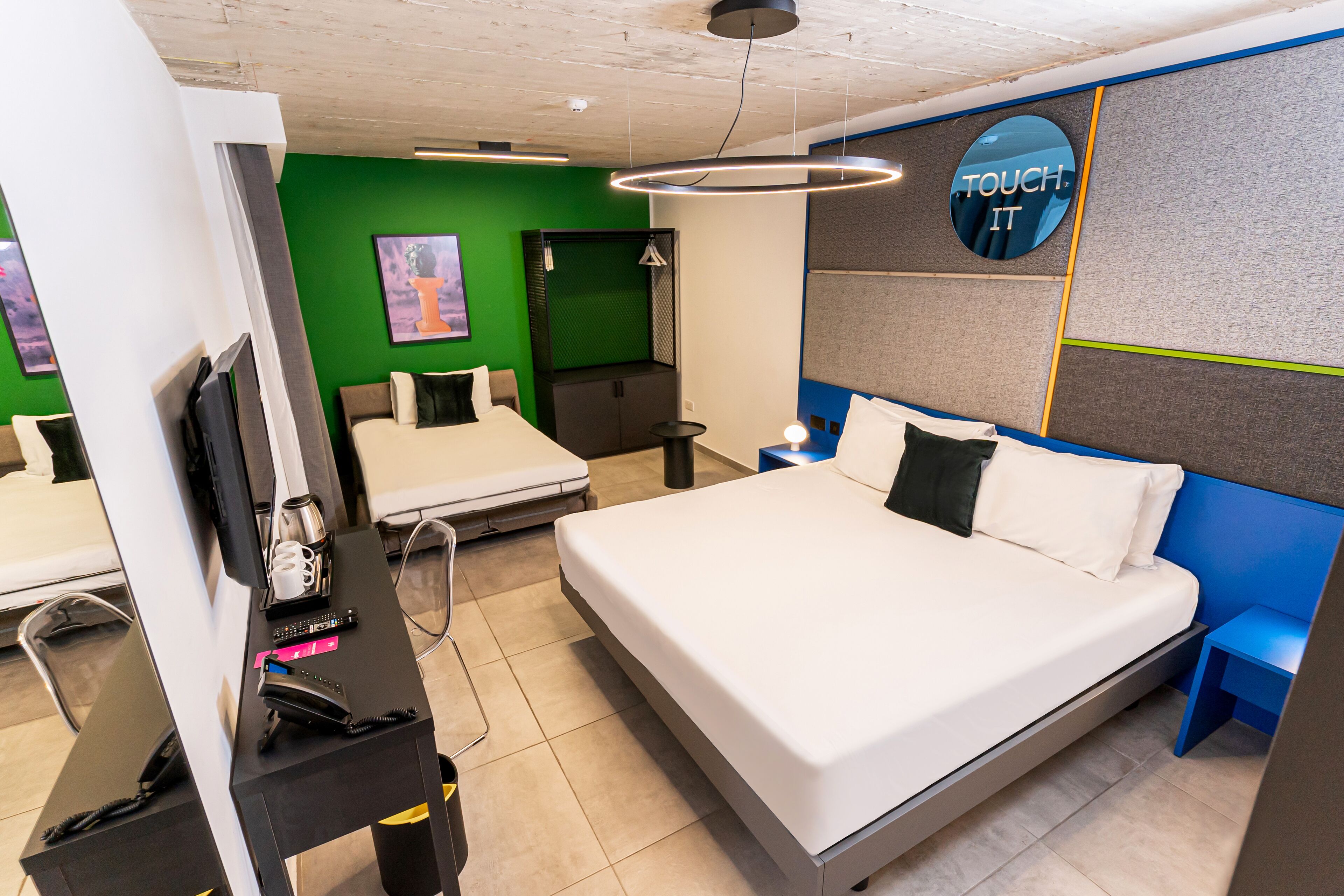 Una habitación de hotel con una cama grande, un escritorio con teléfono, una mesita con lámpara y un cartel en la pared que dice 'TOUCH IT'.
