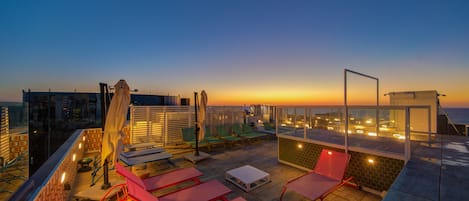 Une terrasse sur le toit, dotée de chaises longues, d’une table et d’une balustrade en verre, offrant une vue sur le paysage urbain au coucher du soleil.