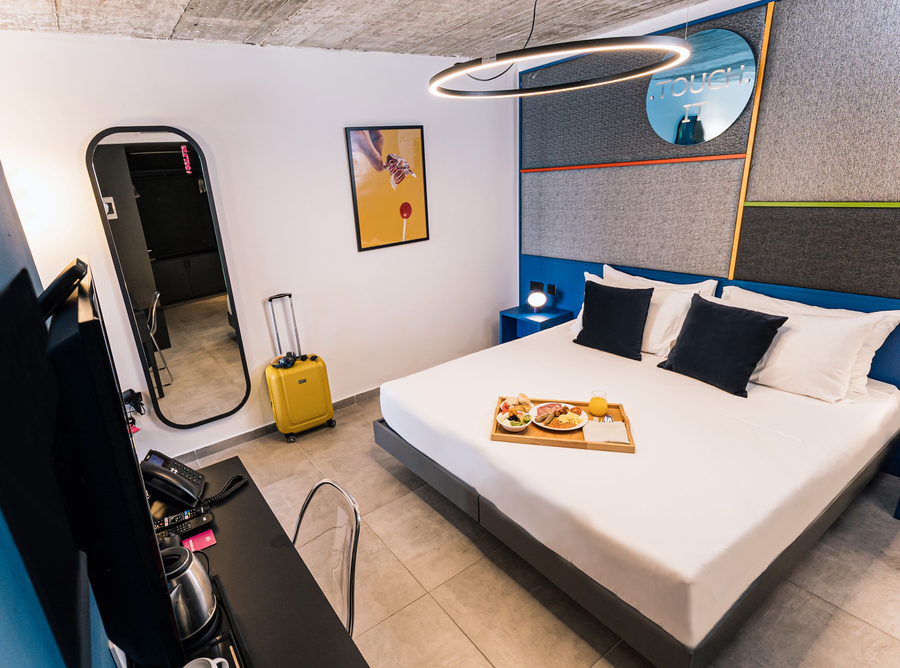 A hotel room with a bed, a bedside table, a mirror, a wall art, a nightstand, and a suitcase.