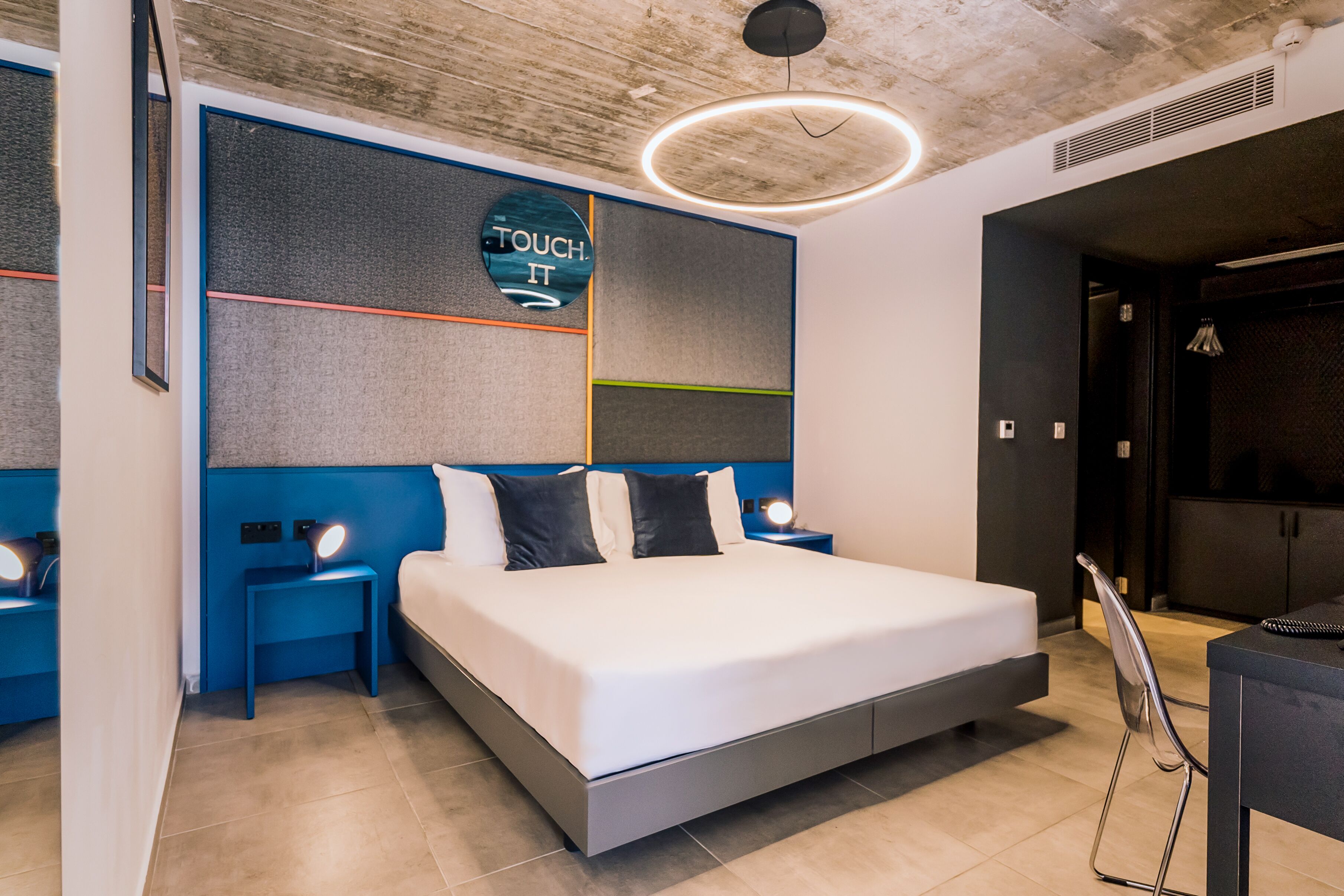Un dormitorio moderno con una cama grande, dos mesitas de noche con lámparas, una lámpara circular fijada a la pared y un panel mural interactivo 'TOUCH IT'.