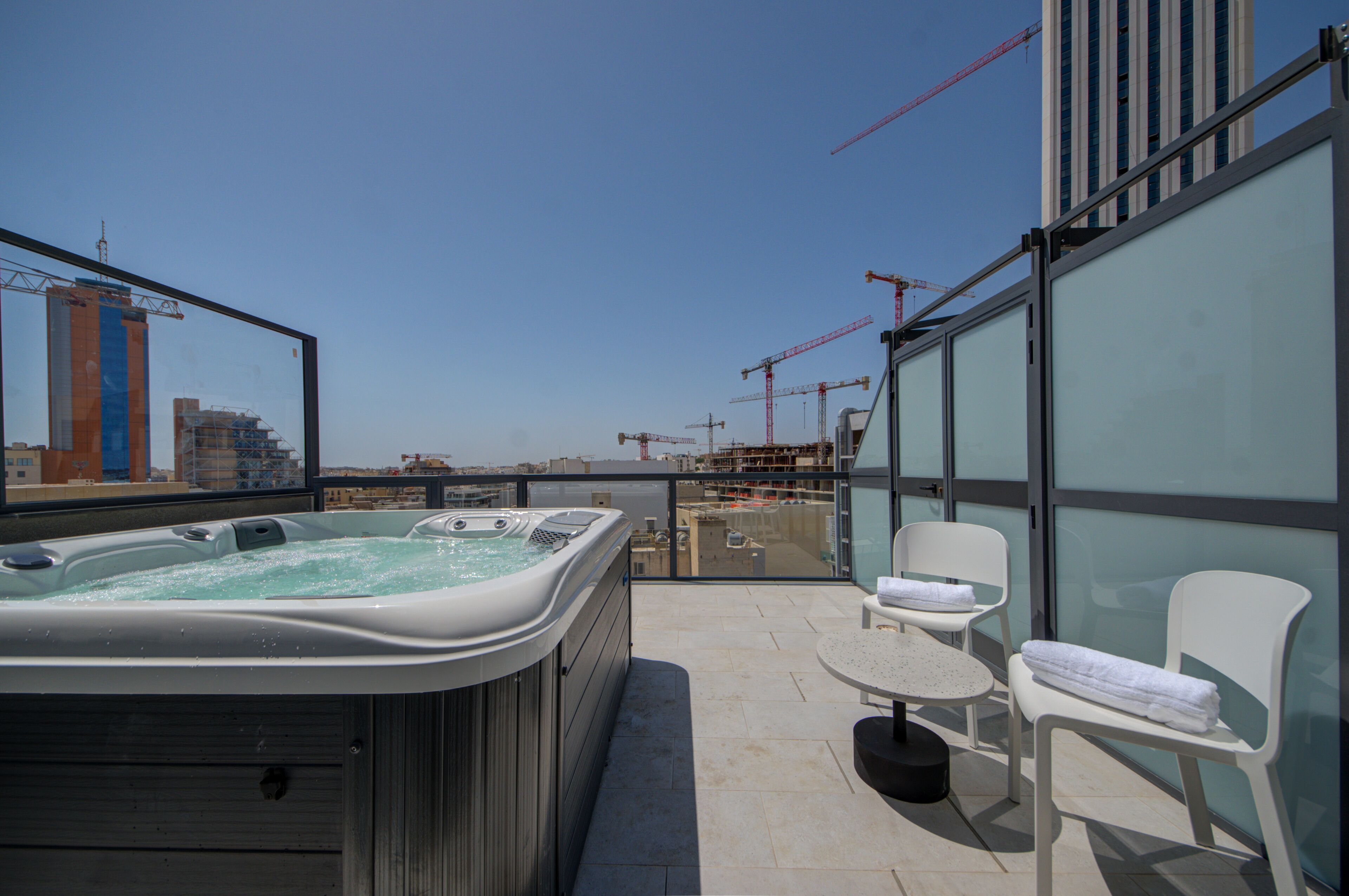 A rooftop terrace with a hot tub, two white chairs, and a glass wall offering a view of the cityscape and construction cranes.