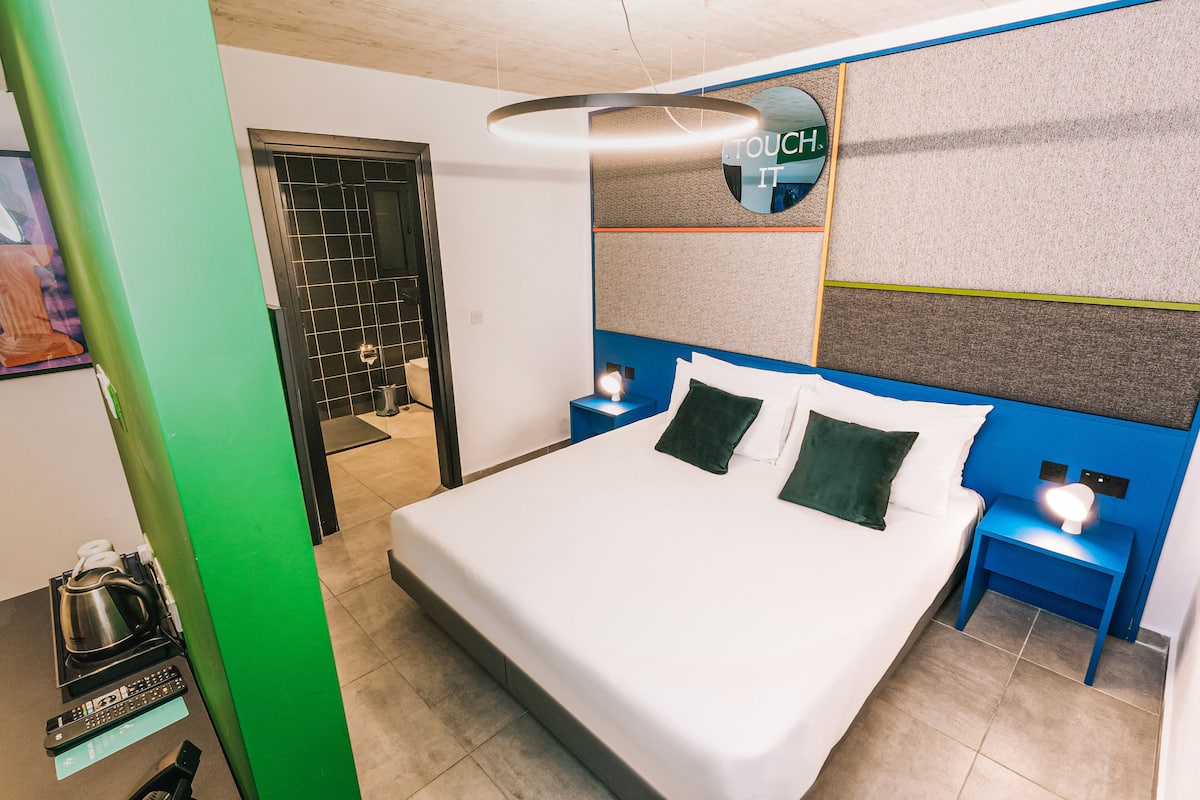 A modern hotel room with a large bed, a nightstand with a lamp, a green wall, and a bathroom visible through an open door.