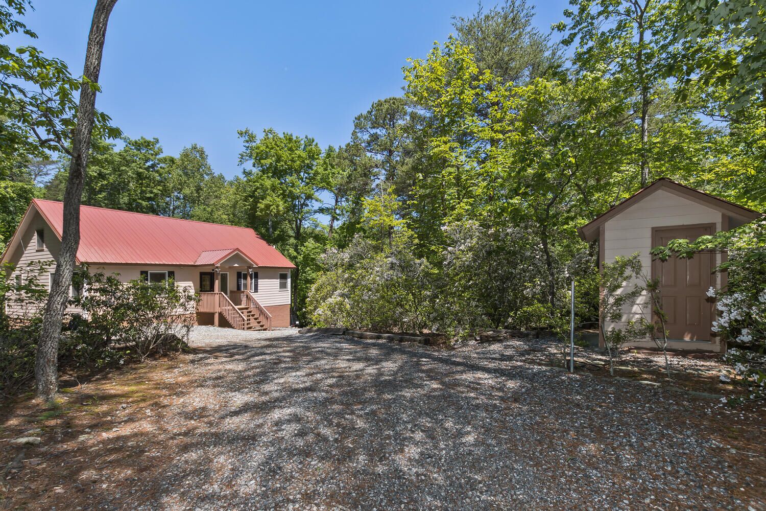 A rustic cabin with a red roof, a wooden porch, and a gravel driveway surrounded by trees.