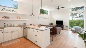 A modern kitchen with white cabinetry, a central island, pendant lighting, and a large window overlooking greenery.