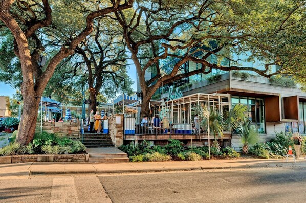 A modern building with a covered outdoor seating area, surrounded by large trees and greenery.
