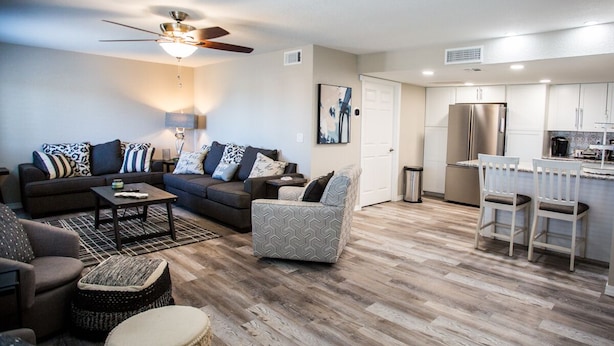 A modern living room with a sofa set, a coffee table, and a patterned ottoman. There is a kitchen area with white cabinets and a stainless steel refrigerator. - Prinstine 4BR Havasu Home — Tons of Boat Parking & Dual AC (Lake Havasu City)