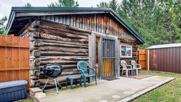 Une cabane rustique avec une façade en bois, une porte d’entrée, une fenêtre et un espace patio aménagé avec des sièges d’extérieur.