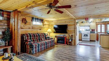 L’intérieur d’un chalet accueillant, avec un plancher en bois, un canapé à carreaux, une cheminée, un téléviseur et un coin cuisine.