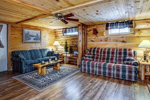 A log cabin interior with a sofa, armchair, and a small table with a lamp.