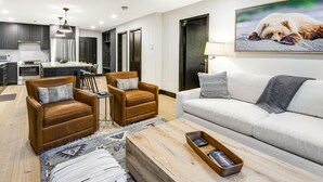 Een moderne woonkamer met een bank, twee fauteuils, een salontafel en een keuken op de achtergrond.