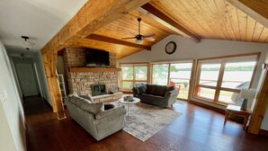 A spacious living room with a stone fireplace, wooden ceiling beams, and large windows offering a view of the outdoors. - Need a summer getaway? Book NorthFork Lakehouse-the ultimate cottage destination (Sebright)