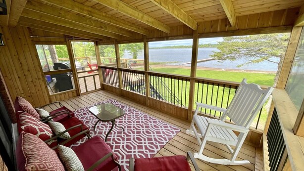 A covered deck with a wooden floor, red and white patterned rug, a glass table, and a white rocking chair. - Need a summer getaway? Book NorthFork Lakehouse-the ultimate cottage destination (Sebright)