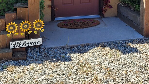 Una puerta marrón con un adorno de lazo, dos cestas de flores colgantes, un felpudo y un letrero que dice 'Welcome'.