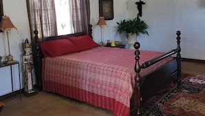 Un dormitorio con cama de madera, ropa de cama roja, alfombra estampada, mesita de noche con lámpara y un cuadro colgado en la pared.