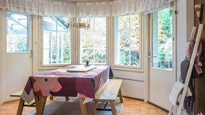 Een kamer met een grote tafel bedekt met een kleurrijke tafelkleed, een bank en een kroonluchter. Er zijn grote ramen met gordijnen, en een schilderij aan de muur.