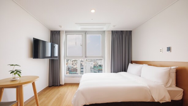 A hotel room with a bed, a TV mounted on the wall, a small table with a plant, and a window with curtains. - KENNY STAY BUSAN GIJIANG - HOTEL KENNY (Busan)